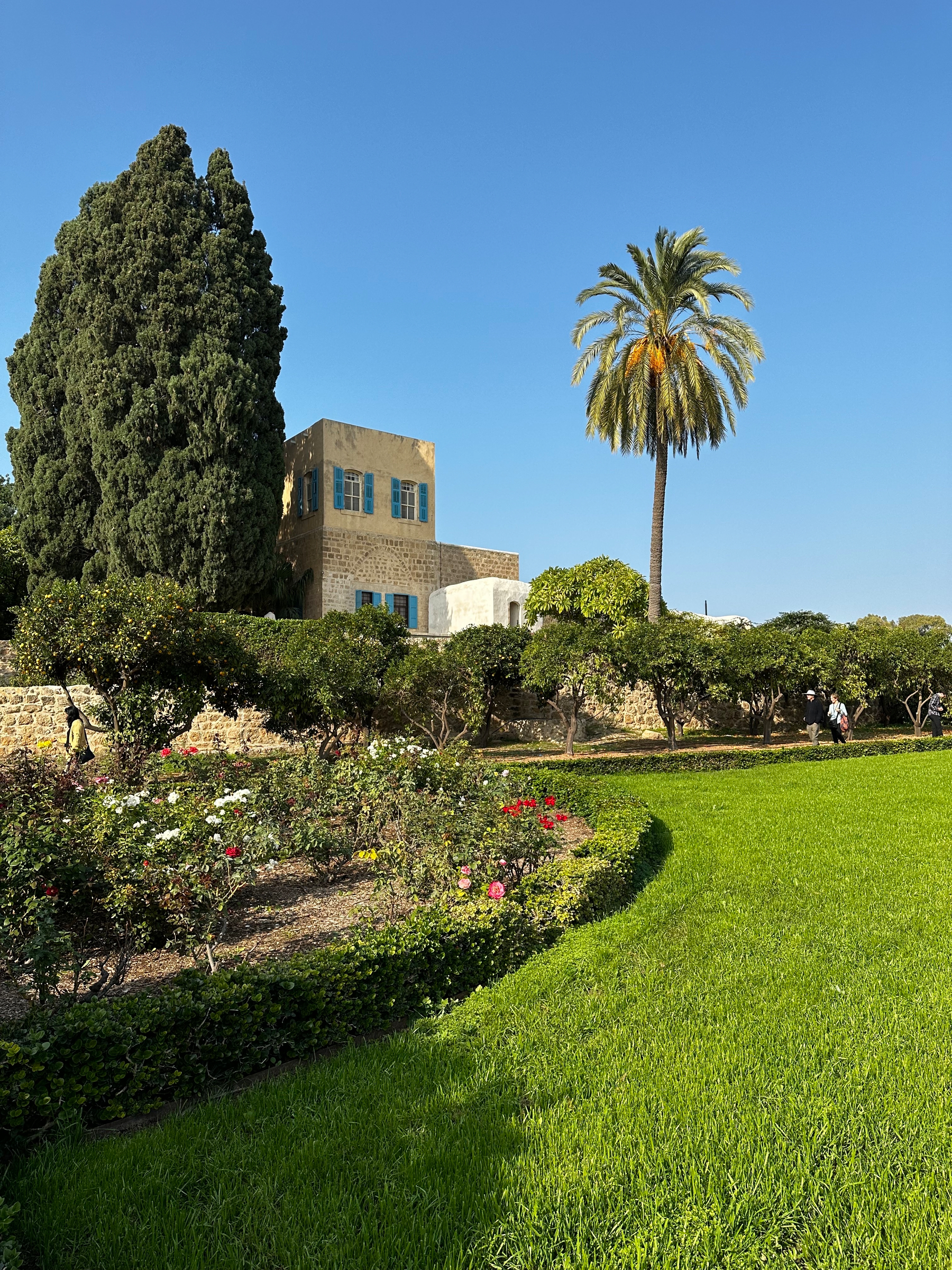 A lush garden with neatly trimmed grass and flower beds is bordered by tall trees and a stone building under a clear blue sky.